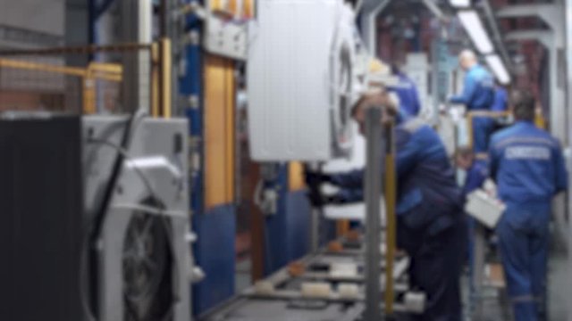 Blurred view of conveyor, masters and workers at work, part assembly process of element washing machine, assembly shop.