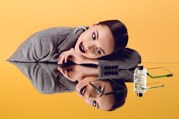 smiling glamorous woman in formal wear with mirror reflection and perfume bottle isolated on yellow