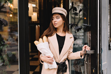 Modern stylish woman wearing beige coat amd cap comes outs from french bakery with baguette ands looks aside with lovely smile. Pretty brunette on the street in a semi-profile, looking confusedly down