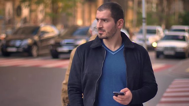 Portrait of caucasian man discontentedly watching into smartphone with blurred walking people on background.
