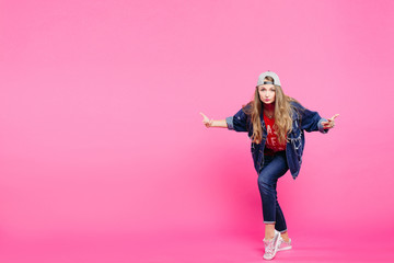 Stylish and beautiful young girl with long, chestnut hair, wearing jeans outfit, cap and sneakers. Fashionable and trendy model standing and posing in studio with pink background.