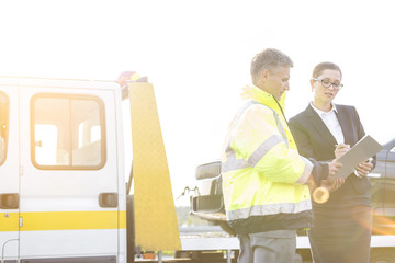 Businesswoman signing report while talking to tow truck driver against sky