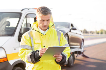 Mature tow truck driver making reports on clipboard against sky