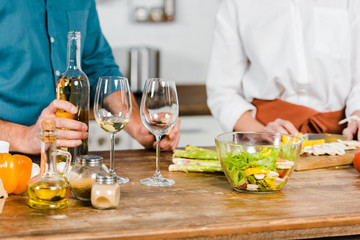 cropped image of mature wife cutting vegetables and husband holding bottle of wine in kitchen