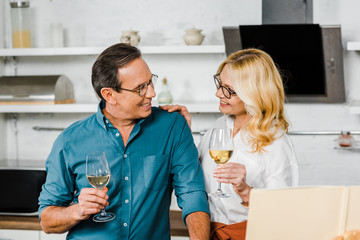 mature wife and husband holding glasses of wine and looking at each other in kitchen