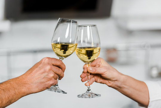 cropped image of wife and husband clinking with glasses of white wine in kitchen