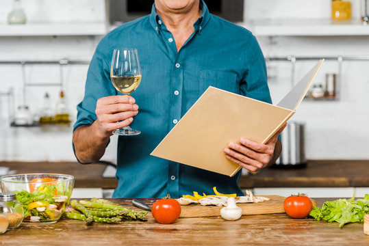 cropped image of mature man holding glass of wine and cookbook in kitchen
