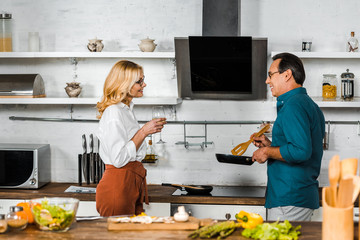 mature wife holding glass of wine and husband cooking at stove in kitchen