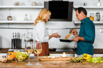 selective focus of mature husband and wife looking at frying pan in kitchen, wineglasses on tabletop
