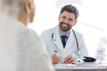 Smiling doctor discussing with mature patient while writing prescription at desk in hospital - obrazy, fototapety, plakaty