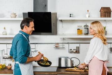 mature wife cutting vegetables and husband cooking on frying pan in kitchen