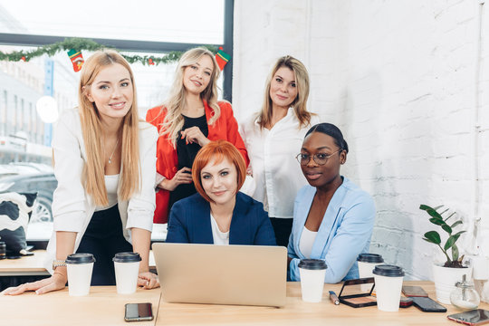 Group of designers working on a project as a team, diverse women gathered together near laptop of red-haired female colleague.