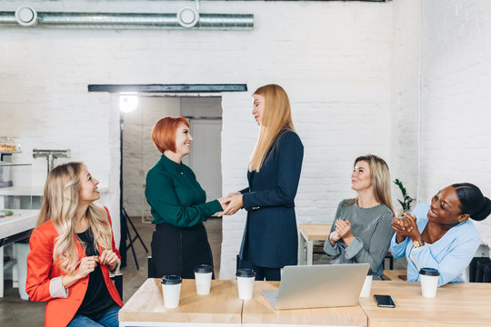 Happy blonde female boss promoting employee to a new position, hand shaking with female co-worker showing respect while team applauding at group meeting, appreciation and employee recognition concept