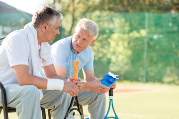 Fototapeta na wymiar Mature male friends talking while sitting with tennis rackets and water bottle on chair at court