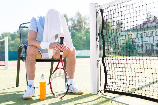Full length of tired mature man with covered head sitting on chair by net at tennis court on sunny day