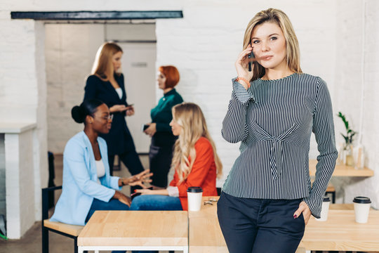 Caucasian mid-aged female manager, casual dressed, using smartphone in well-lit white office interior with her co-workers on background