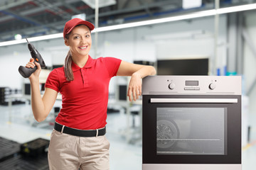 Repairwoman in a red t-shirt and a red cap with a drill and an oven in a factory