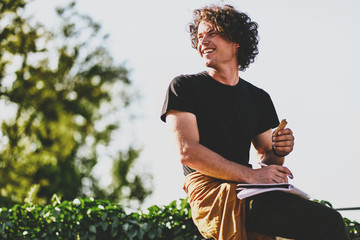 Successful smiling male student with curly hair, smiling and wearing black t-shirt sitting on the...