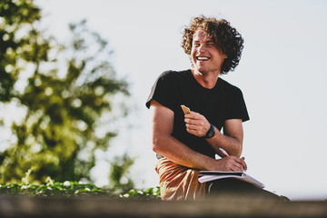 Confident happy guy with curly hair, smiling and wearing black t-shirt sitting on the street and preparing for exams. Freelancer businessman planning the day sitting on the city street