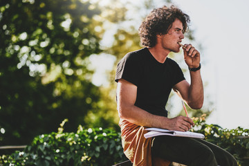 Side view portrait of good-looking man student with curly hair, wearing black t-shirt, eating snack and preparing for exam. Freelancer businessman planning the day sitting on the city street
