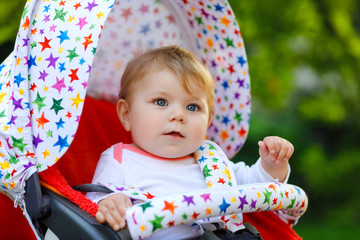 Fototapeta na wymiar Cute healthy little beautiful baby girl sitting in the pram or stroller and waiting for mom. Happy smiling child with blue eyes. With green tree background. Baby daughter going for a walk with family