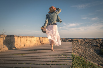 Woman walks barefoot on the floor of wooden planks facing the ocean, long white skirt holds back in...