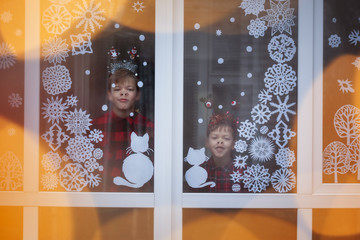 Two adorable brothers are looking through the window. There is decoration snowflakes from paper on the window.