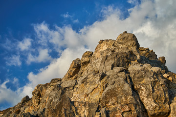 Coastal rocky peak of a cliff with white clouds on a blue sky in summer in Greece.