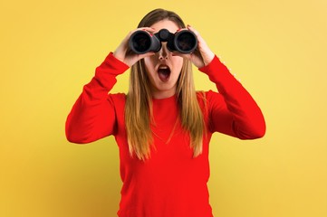 Young blonde woman with binoculars on yellow background