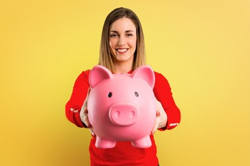 Obraz premium Happy Young blonde woman holding a piggybank on yellow background