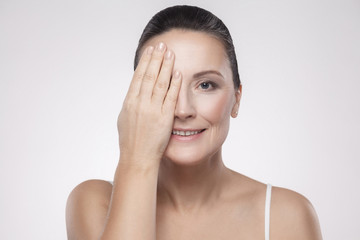 Naklejka premium Closeup portrait of happy toothy smiling charming, beautiful, pretty middle aged woman with perfect face skin hiding one eye with hand isolated on grey background. Indoor, studio shot, copy space
