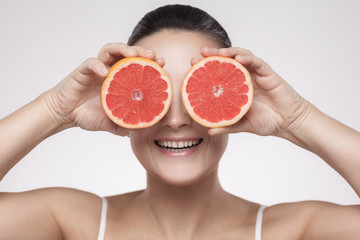 Closeup portrait of happy woman with perfect skin smiling after cream, balm, mask, lotion, holding half of grapefruit and covering her eye, isolated on grey background. Indoor, studio shot,copy space