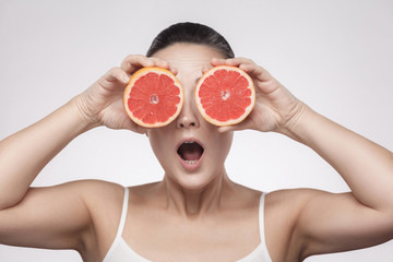 Closeup portrait of suprized woman with perfect skin smiling after cream, balm, mask, lotion, holding half of grapefruit and covering her eye, isolated on grey background. Indoor, studio shot.