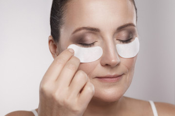 Closeup portrait of beautiful middle aged woman with perfect skin using white hydrogel patches with lifting anti-wrinkle collagen effect, gray background, indoor, studio shot, copy space, isolated