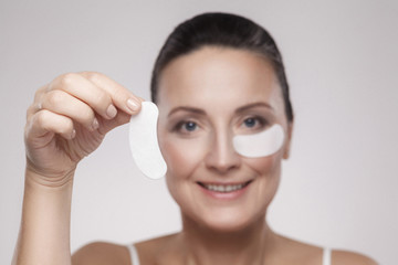 Patch Under Eyes. Beautiful middle aged woman with perfect skin showing white hydrogel patches with lifting anti-wrinkle collagen effect, gray background, indoor, studio shot, copy space, isolated