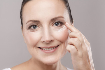 Middle aged woman with perfect face skin applying cosmetic cream on skin near eyes and looking at camera with toothy smile isolated on grey background. Skin care concept,copy space,studio shot, indoor