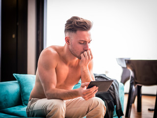 Obraz na płótnie Canvas Shirtless young athletic man holding ebook reader or tablet PC and reading while sitting on couch