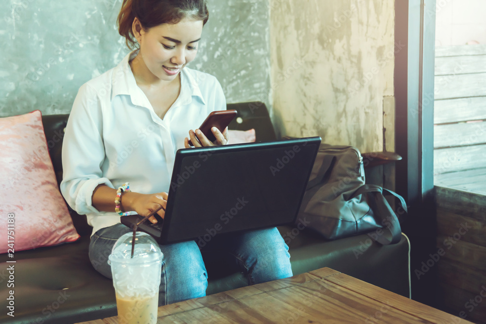 Wall mural asian woman sitting relax on the sofa holding the mobile phone and is working with a notebook. at a 