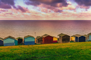 Moody cloudy weather creating patches of sunlight between the clouds on the sea in Tankerton, Whitstable in Kent.  Rows of wooden beach huts line the coastline with green sloping banks behind them