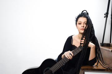Cute hipster girl wearing a black top and blue jeans holding a guitar posing on a staircase against a white background.