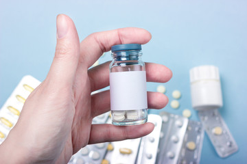 Female hand holding a glass bottle of medicine and a blank label on a background of drugs, tablets, pills and capsules. With blank placer space for text or design