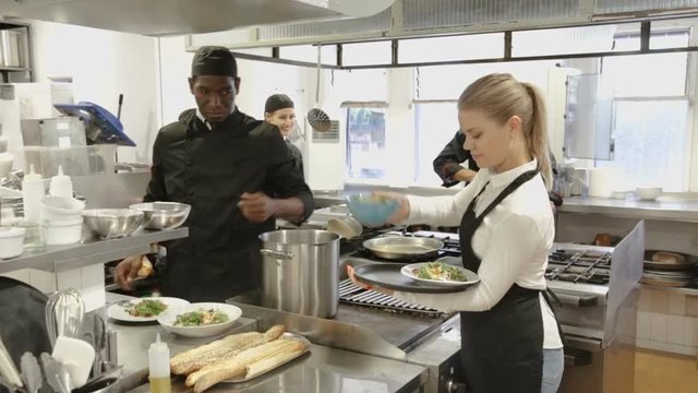 Attractive young waitress taking prepared dishes from chef in kitchen of modern restaurant 