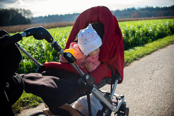 Obraz premium Adorable baby girl outside in red stroller in green fields on a road, sunny day.