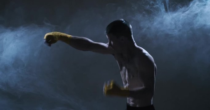 Muscular kickboxing man in yellow gloves shows the different movements and strikes in the studio. Fog in the background