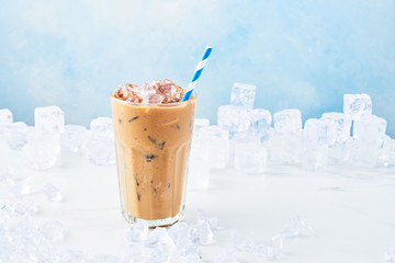 Summer drink ice coffee with cream in a tall glass with straw surrounded by ice on white marble table over blue background. Selective focus, copy space for text.