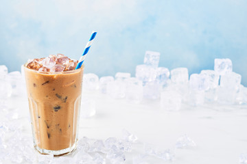 Summer drink ice coffee with cream in a tall glass with straw surrounded by ice on white marble table over blue background. Selective focus, copy space for text. Horizontal.