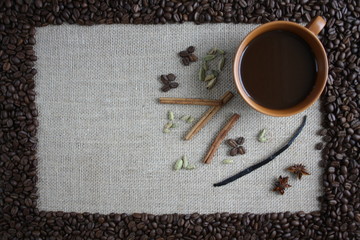 Obramowanie z ziaren kawy, wewnątrz na tkaninie z juty filiżanka czarnej kawy i przyprawy: ziarna kardamonu, laski cynamonu, wanilia i anyż