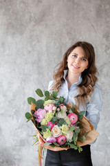 Young friendly smiling female floral artist looking at the camera, holds a luxurious bouquet in his hands. Professional florist making a bouquet of pink Ranunculus and peonies in a flower shop.