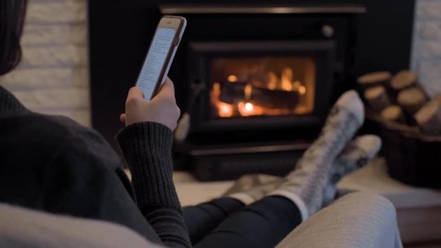 Low light setting of woman by wood burning fire place scrolling on cell phone through social media relaxing and enjoying her comfortable day off  while wearing winter fall season socks