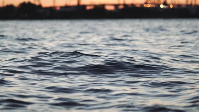 Maritime city port. Slow motion quiet rocking of the sea surface. The ships and cranes on a background of bright orange sunset. Severe ripples. Wave texture. Blue sea water with sky reflection